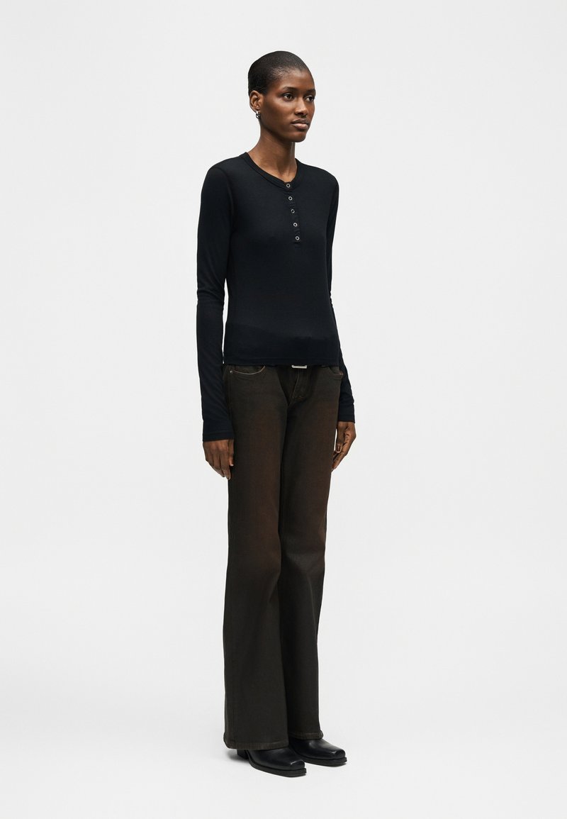 Woman standing wearing a black long-sleeve henley top, dark wide-leg pants, and black shoes against a plain light background.
