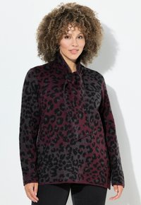 Frau mit lockigem Haar, die einen burgunderroten und schwarzen Leopardenmuster-Pullover mit einer Schleife am Hals trägt, steht vor einem weißen Hintergrund.