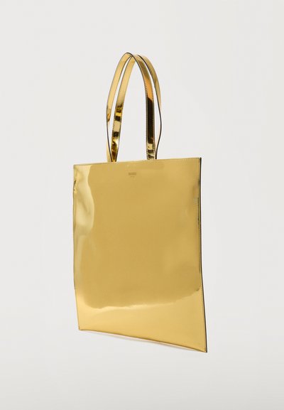 Λαμπερή μεταλλική χρυσή τσάντα tote με δύο μακριές λαβές, λεία επιφάνεια και ορθογώνιο σχήμα κρεμασμένη σε απλό φόντο.