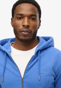 Sudadera de cremallera azul claro con capucha ajustable, textura suave y logo bordado en el pecho. Llevada sobre una camiseta blanca.