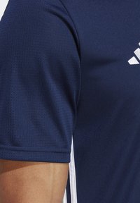 Marineblauw sportshirt van ademende stof, met een textuurpatroon en wit merkembleem op de borst, en contrasterende zijstrepen.