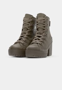 Khaki platform ankle boots with flat laces, round toes, and a textured rubber sole, featuring a logo patch on the side and metal eyelets.