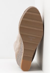 Semelle d'une chaussure compensée beige avec semelle texturée, taille 37, fabriquée en Espagne, positionnée contre un mur blanc d'angle.