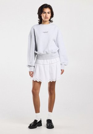 Young woman standing wearing a light gray sweatshirt, white scalloped hem skirt, white socks, and black loafers on a white background.
