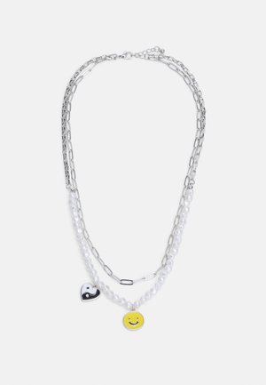 Collier à double chaîne en argent avec des perles blanches, comportant un pendentif cœur yin-yang noir et blanc ainsi qu'un pendentif visage souriant jaune.
