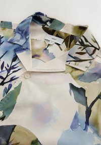 Col et partie haute d'une chemise boutonnée avec un motif floral abstrait en aquarelle dans des tons bleus, verts et beiges, avec une étiquette de taille moyenne.