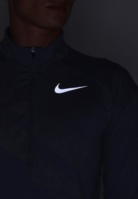 Nike zip-up jacka i mörk marinblått tyg, med ett strukturerat mönster och en framträdande vit logotyp på bröstområdet.