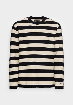 Chemise en tricot à manches longues avec larges rayures horizontales noires et blanches, col rond, et petit texte brodé sur le poignet de la manche gauche.