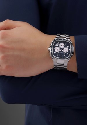 Silver stainless steel wristwatch with a black textured dial, three sub-dials, and a curved rectangular case. Attached to a linked bracelet.