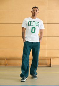 Hvid T-shirt med grøn "CELTICS" tryk og nummer "0", parret med mørkegrønne vidde bukser og brune og hvide sneakers. Simpelt design.