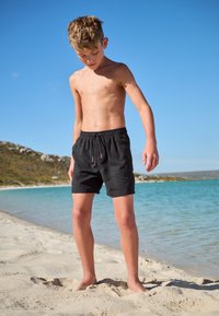 Sorte badeshorts med elastisk talje og snøre, der har sidelommer. Omgivelserne inkluderer sandstrand og vand.