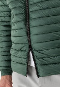 Green padded jacket featuring quilted texture, front zipper, and ribbed cuffs, designed for warmth and comfort. Accents include black zipper.