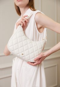 White quilted leather handbag with a curved shape, featuring a gold accent and a top handle. The texture is smooth with diamond patterns.