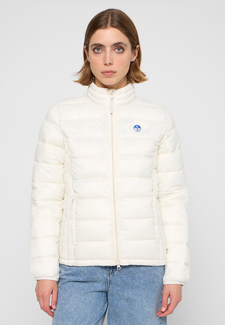 Giacca puffer crema con colletto alto, cuciture orizzontali e un piccolo logo blu sul lato sinistro del petto. Chiusura con zip e tasche laterali.