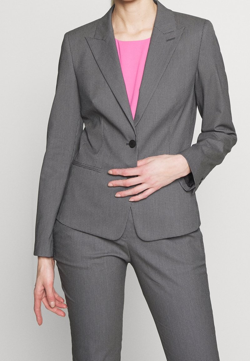 Costume gris sur mesure avec un motif texturé. Comprend un blazer à bouton unique et un pantalon slim, accompagné d'un haut rose vif.