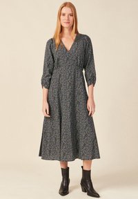 Schwarzes Midi-Kleid mit lila Blumenmuster, V-Ausschnitt, drei Viertelärmel und gerafftem Taillenbereich, kombiniert mit schwarzen Knöchelschuhen.