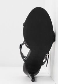Sandale noire à talons hauts avec bride et talon fin, vue de la semelle sur fond blanc.