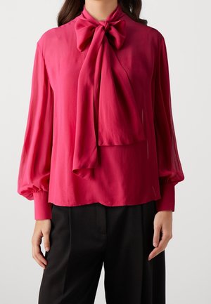 Femme portant un chemisier rose vif à manches longues avec un grand nœud au col, associé à un pantalon noir.