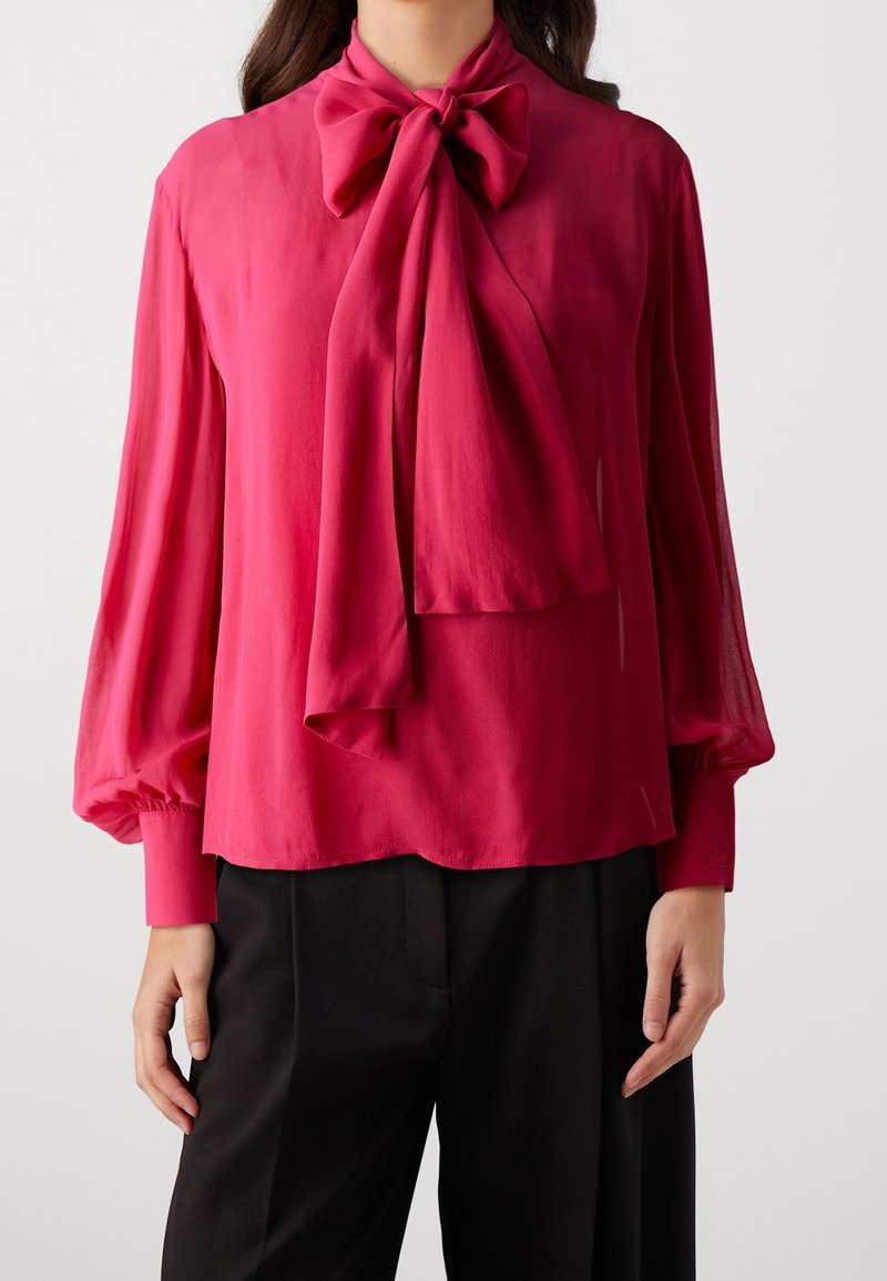 Femme portant un chemisier rose vif à manches longues avec un grand nœud au col, associé à un pantalon noir.