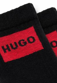 Sorte sokker med et rødt bånd, der har ordet "HUGO" i sorte bogstaver. Materialet ser blødt ud med en tekstureret overflade på stoffet.