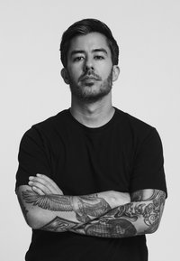 Porträtt av en person som bär en svart t-shirt. Märkbara tatueringar av fåglar och intrikata mönster täcker deras armar. Neutral bakgrund.