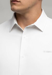 Biała koszula z kołnierzykiem, zapinana na guziki, o gładkiej fakturze i subtelnym haftem 'Lee Cooper' na klatce piersiowej. Klasyczny krój.