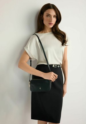 Kobieta o długich brązowych włosach, ubrana w białą bluzkę i czarną spódnicę, trzymająca czarną torbę na ramię z cienkim paskiem na jednolitym tle.