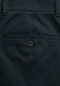 Pantaloni din țesătură albastru închis, cu un model texturat, o ușoară strălucire și un buzunar posterior cu detaliu dintr-un buton negru.