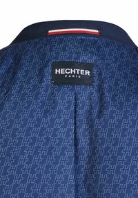Granatowa tkanina z powtarzającym się wzorem litery "H", czarna metka z napisem "HECHTER PARIS" oraz trójkolorowa etykieta w kolorach czerwonym, białym i niebieskim na kołnierzu.