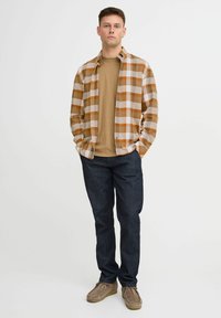 Karierter Hemd in Tan- und Brauntönen über einem braunen T-Shirt, kombiniert mit dunklen Jeans und hellbraunen Schuhen. Neutrale Hintergrund.