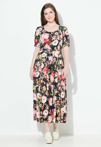 Frau, die in einem schwarzen, blumigen Midikleid mit Knöpfen an der Vorderseite und weißen Clogs vor einem weißen Hintergrund steht.