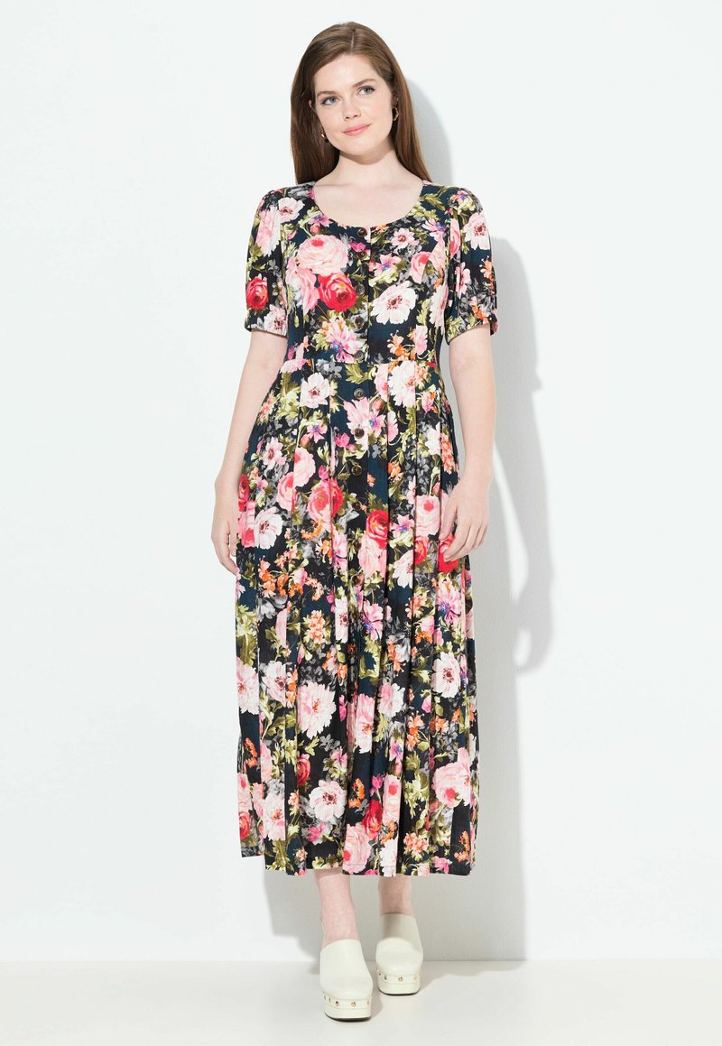 Frau, die in einem schwarzen, blumigen Midikleid mit Knöpfen an der Vorderseite und weißen Clogs vor einem weißen Hintergrund steht.