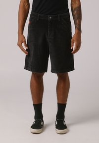 Schwarze Cordshorts mit vertikalen Rippstrukturen, ausgestattet mit einem Frontknopfverschluss, Gürtelschlaufen und seitlichen Taschen. Getragen mit schwarzen Socken und Schuhen.