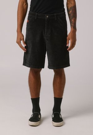 Shorts en velours côtelé noir avec des côtes verticales, dotés d'une fermeture à bouton à l'avant, de passes-belt et de poches latérales. Portés avec des chaussettes et des chaussures noires.