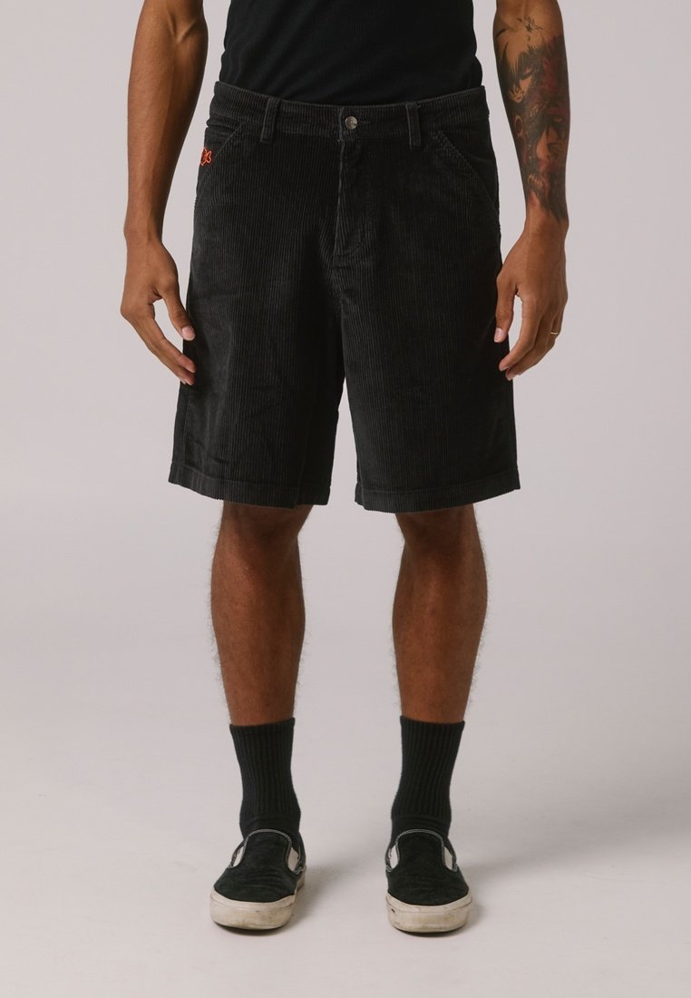 Schwarze Cordshorts mit vertikalen Rippstrukturen, ausgestattet mit einem Frontknopfverschluss, Gürtelschlaufen und seitlichen Taschen. Getragen mit schwarzen Socken und Schuhen.