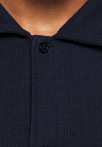 Polo de punto azul marino con una tela texturada, que presenta un cuello y un solo botón negro en la tapeta para el cierre.