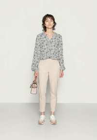 Mulher em pé a usar uma blusa de manga longa estampada, calças cropped bege, ténis brancos e a segurar uma pequena mochila ao lado.