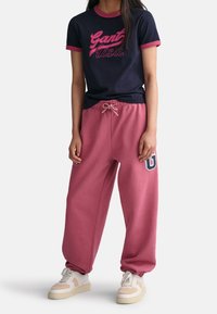 Camiseta navy con ribete rosa y texto "Gant USA", combinada con pantalones de chándal rosas con cordón y detalle de logo, llevados con zapatillas blancas.