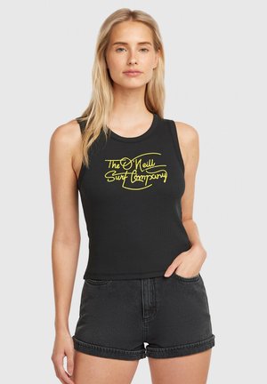 Čierny crop top bez rukávov s rebrovanou textúrou a žltou potlačou loga, v kombinácii s čiernymi džínsovými šortkami s vyhrnutými okrajmi.