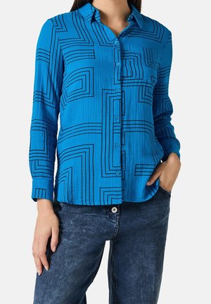 Femme portant une chemise boutonnée à manches longues bleu vif avec des lignes géométriques noires et un jean bleu foncé, une main dans la poche.