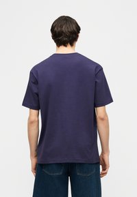 Άνδρας με σκούρα μαλλιά που φοράει μονόχρωμο μπλε σκούρο T-shirt με κοντά μανίκια και μπλε τζιν παντελόνι, γυρισμένος προς τα πίσω σε ανοιχτό γκρι φόντο.