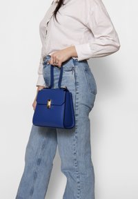 Blauwe leren handtas met gouden accenten, rechthoekige vorm en dubbele handvatten. Draagt bij lichte spijkerbroek en een witte overhemd.