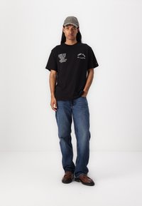 Sort bomulds t-shirt med hvide tekstgrafikker, parret med bløde blå jeans. Modellen bærer en grå kasket og brune sko.