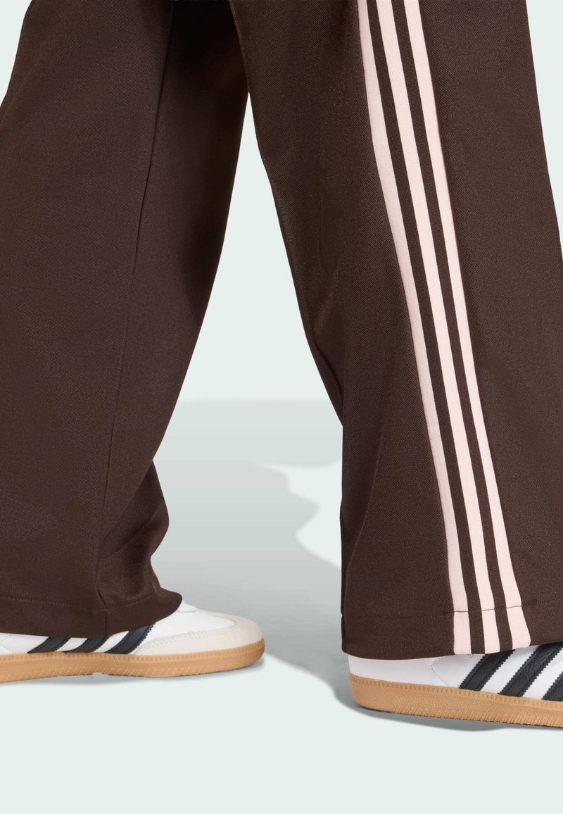 Brown track pants with light pink stripes and white athletic shoes featuring black stripes and a textured gum sole.