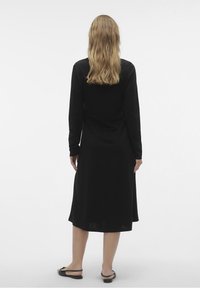 Mujer con cabello largo y rubio, vistiendo un vestido midi negro de manga larga y sandalias planas negras, de pie de espaldas sobre un fondo blanco.