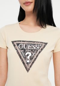 Camiseta de algodón beige con un gran logo triangular negro y plateado que incluye el texto "GUESS" y un signo de interrogación, sobre un fondo texturizado.