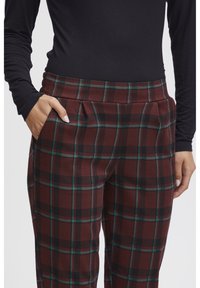Pantaloni a quadri marroni con accenti verdi e neri, una piega in vita e tasche laterali. Il materiale sembra morbido con una finitura testurizzata.