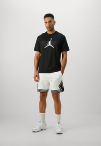 Sort T-shirt med et hvidt grafisk logo, parret med hvide mesh shorts med sorte accenter og striber, suppleret af hvide sneakers.