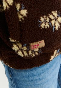 Hnědý fleece oděv s krémovými a modrými vzory, se strukturou povrchu. Obsahuje textilní etiketu "Cítím se šťastný".