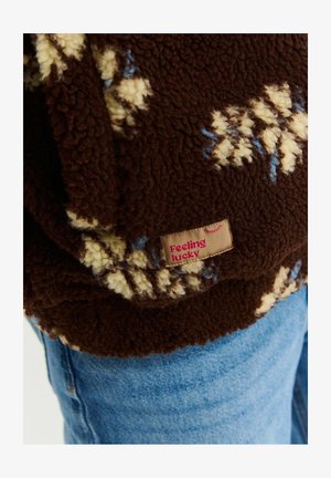 Hnědý fleece oděv s krémovými a modrými vzory, se strukturou povrchu. Obsahuje textilní etiketu "Cítím se šťastný".